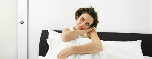 Portrait of smiling, happy young woman with sensual face, relaxing in bed, waking up, sitting with dreamy expression, resting at home photo