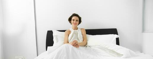 Image of smiling candid female model, lying in bed under white sheets, looking happy at camera, concept of morning, waking up early photo