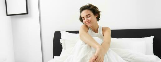 Portrait of cute smiling woman, sitting in bed, resting at home, lying under white linen sheets, waking up in morning with happy, pleased face expression photo