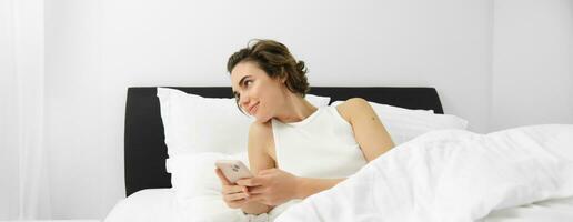 Portrait of woman in bed, lying on white sheets, using smartphone, holding mobile phone photo