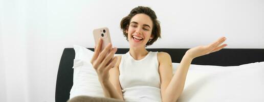 Smiling friendly young woman chatting on smartphone app, talking via video call app on mobile phone, lying in bedroom, resting in bed and communicating with someone photo