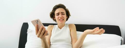 Portrait of woman with confused face, laying in bed with smartphone, chatting on mobile phone app, video call, shrugging shoulders at some nonsense, feeling upset by something she sees online photo