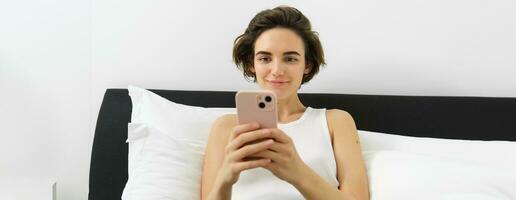 Portrait of smiling brunette woman resting in her bedroom, using mobile phone and lying in bed in her home outfit, messaging, sending message on smartphone app photo