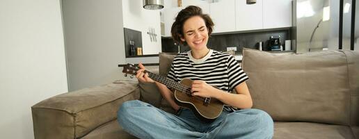 joven mujer jugando ukelele con pasión, canto canción, rasgueo instrumentos de cuerda, sentado en sofá en vivo habitación a hogar foto