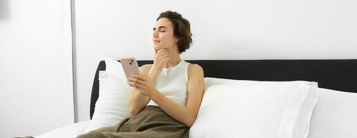 Portrait of young woman with eyes closed and pleased smile, holding smartphone, lying and resting in bed, using mobile phone photo