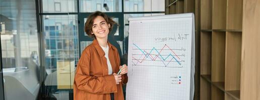 Portrait of successful businesswoman, modern manager in office, standing near board with diagram, giving presentation, giving speech in conference room photo