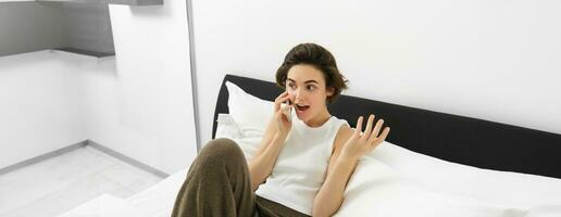 Chatty young woman laughing, talking on phone, calling friend, lying in bed and having conversation with someone, resting in bedroom photo