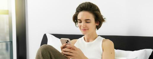 Portrait of young beautiful brunette woman, sitting on bed with smartphone, using mobile phone app, sending message and smiling, reading on telephone application in bedroom photo