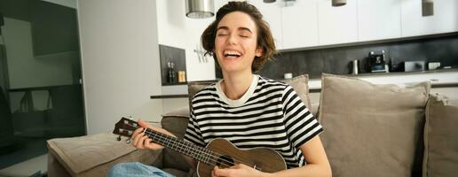 Young happy woman sitting on sofa and playing ukulele, singing and enjoying learning new musical instrument photo