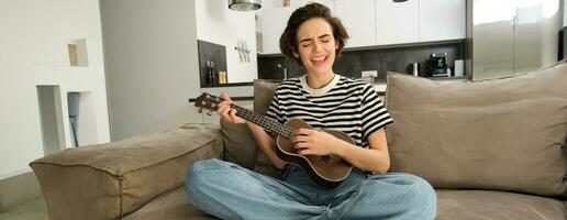 joven contento mujer sentado en sofá y jugando ukelele, canto y disfrutando aprendizaje nuevo musical instrumento foto