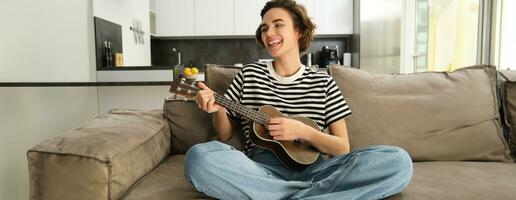 alegre joven mujer con ukelele, jugando musical instrumento, participación pequeño guitarra y cantando, sentado en sofá en cruzado piernas, descansando en vivo habitación foto