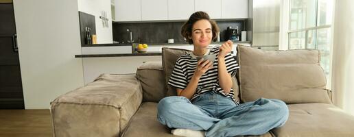 Portrait of smiling cute woman eating cereals for breakfast with satisfaction, sitting on sofa in living room and having a snack photo