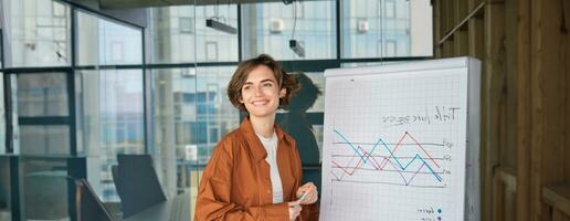 Portrait of young woman manager in office, standing near glass wall in conference wall, showing diagram, company presentation photo