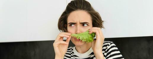 Portrait of woman frowning and grimacing, smelling fresh lettuce leaf with displeased face, standing in the kitchen, dislike vegetables photo