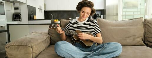 retrato de linda joven mujer sentado en sofá, aprende cómo a jugar ukelele, participación su música instrumento, cosecha acordes, descansando en vivo habitación foto