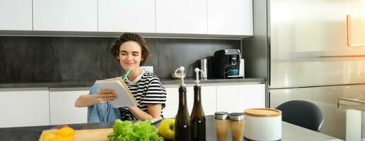 retrato de joven mujer mirando a Cocinando ingredientes en cocina mostrador y haciendo notas, escritura abajo recetas, pensando de comida para cena, preparando vegetariano comida foto