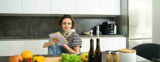 retrato de joven sonriente mujer en cocina, participación computadora portátil, haciendo notas para receta, escritura tienda de comestibles lista, Cocinando ensalada, sentado cerca vegetales y el cortar tablero foto