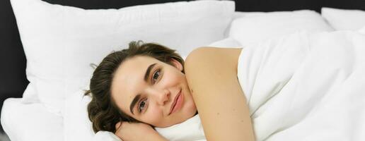 Close up portrait of sensual, beautiful young woman lying in bed on soft white pillow, gazing at camera with happy, dreamy smile photo