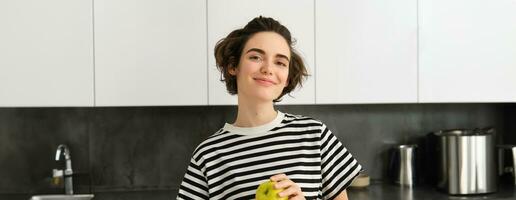 retrato de hermoso, sonriente joven mujer, participación un manzana, comiendo Fruta en el cocina, mirando contento. concepto de sano dieta y estilo de vida foto