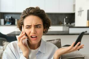 Image of woman with confused, annoyed face, talking on mobile phone while sitting at home on sofa, shrugging and looking bothered photo