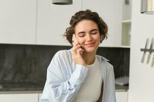 cerca arriba retrato de hermosa europeo niña con teléfono inteligente, sentado a hogar y hablando en móvil teléfono, vocación alguien y sonriente foto