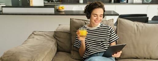 Portrait of woman in headphones, sitting on sofa with tablet and drinking orange juice, watching tv show on her gadget, using social media application, spending time at home photo