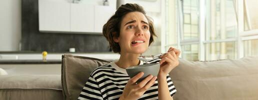 Close up portrait of female model, girl eating breakfast and watching intense scene in movie, staring at tv screen in living room, holding bowl of cereals with milk photo