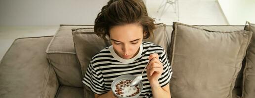 Upper angle shot of happy, cute young woman on sofa, eating bowl of cereals with milk and smiling, enjoying her breakfast photo