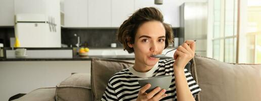 retrato de contento morena mujer sentado en sofá y disfrutando su desayuno, comiendo cereales, sano Granola en bol, gasto hora en vivo habitación foto