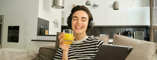 Close up portrait of young brunette woman in living room, watching funny video or tv series on digital tablet, drinking orange juice, sitting on sofa and laughing photo