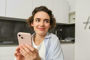 Portrait of modern brunette woman holding mobile phone and smiling. Girl sits in kitchen at home with smartphone, using app to order delivery, does online shopping photo