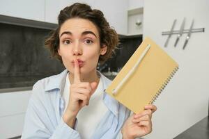 Young woman holds her diary, shows secret, hush gesture, presses finger to lips, puts her thoughts in writing photo