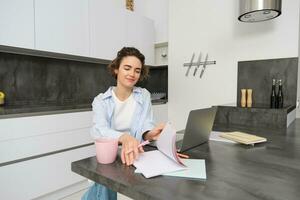 joven determinado mujer, voltea mediante paginas en su libro de trabajo, estudios en remoto, en línea aprendizaje desde hogar, se sienta en frente de ordenador portátil foto