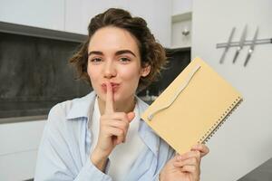 Young woman holds her diary, shows secret, hush gesture, presses finger to lips, puts her thoughts in writing photo