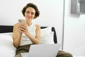 Image of stylish modern woman, sitting on bed, resting at home with laptop and smartphone, sending message, reading notification on mobile phone photo
