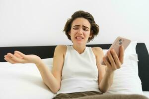 Portrait of woman with confused face, laying in bed with smartphone, chatting on mobile phone app, video call, shrugging shoulders at some nonsense, feeling upset by something she sees online photo