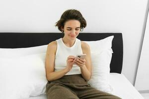 Portrait of young beautiful brunette woman, sitting on bed with smartphone, using mobile phone app, sending message and smiling, reading on telephone application in bedroom photo