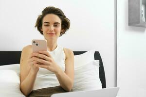 Image of stylish modern woman, sitting on bed, resting at home with laptop and smartphone, sending message, reading notification on mobile phone photo