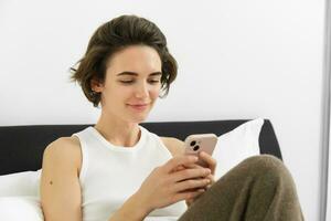 Portrait of young beautiful brunette woman, sitting on bed with smartphone, using mobile phone app, sending message and smiling, reading on telephone application in bedroom photo