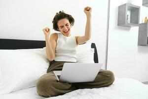 Portrait of happy woman rejoicing, sitting with computer and doing winner dance, has laptop on her laps, raising hands up and celebrating, triumphing with joy photo