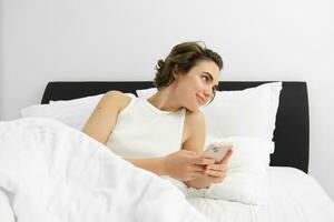 Portrait of woman in bed, lying on white sheets, using smartphone, holding mobile phone photo