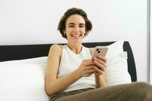 Portrait of smiling brunette woman resting in her bedroom, using mobile phone and lying in bed in her home outfit, messaging, sending message on smartphone app photo