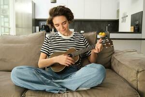 estilo de vida y aficiones concepto. joven sonriente mujer en sofá, jugando ukelele, canto y aprendizaje rasgueo modelo para favorito canción foto