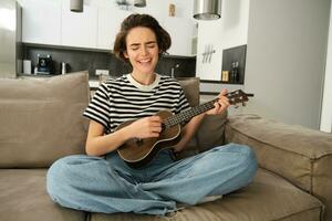 retrato de linda joven mujer, niña con ukelele, jugando y canto favorito canción mientras sentado en vivo habitación en sofá, rasgueo instrumentos de cuerda foto