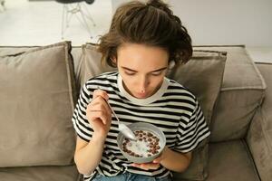 Upper angle shot of happy, cute young woman on sofa, eating bowl of cereals with milk and smiling, enjoying her breakfast photo