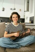 Vertical shot of young musician, woman learns how to play ukulele, sits on sofa with crossed legs and smiling at camera photo