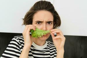 Frowning silly woman, holding lettuce leaf near face and frowning at camera, making pouting face, dislike eating vegetables photo