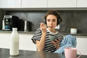 Cute girl eats her breakfast, holding bowl of cereals with milk, drinks coffee and listens to music in wireless headphones, using earphones while having her morning meal photo