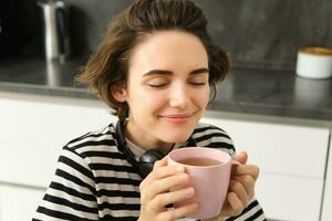 cerca arriba retrato de mujer con cerrado ojos y satisfecho sonrisa, bebidas calentar té con satisfacción, sentado en acogedor cocina foto