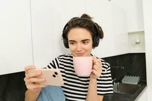 Smiling young woman watching tv series or video blog on smartphone app, drinking tea in the kitchen, looking at mobile phone with interest photo